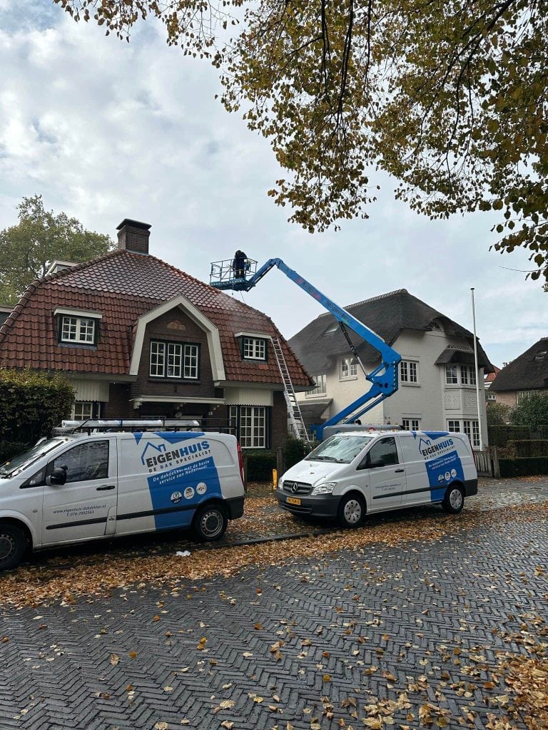 Dakdekker inspecteert woonhuisdak vanuit hoogwerker, twee Eigenhuis Dak specialist busjes geparkeerd