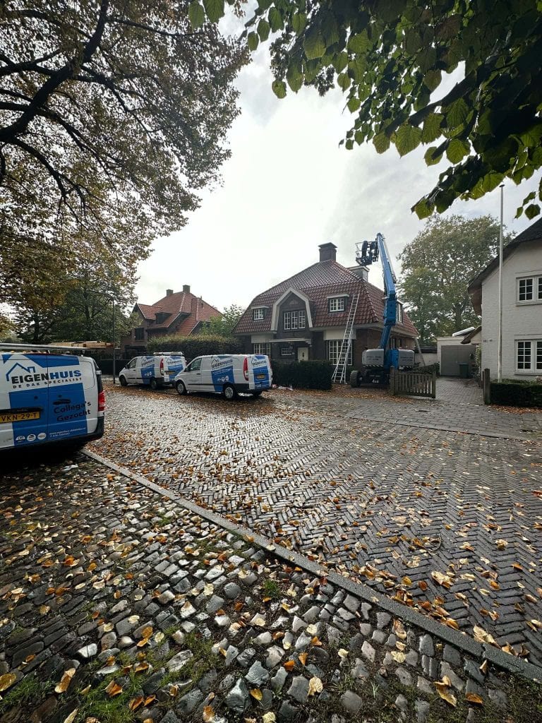 Dakrenovatie bij woning: natte herfstklinkerstraat met Eigenhuis dakspecialistbusjes en hoogwerker